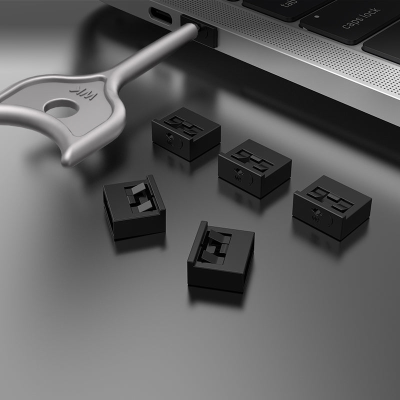 Five small black rectangular port plugs on a matte black surface beside a silver key tool and laptop edge.